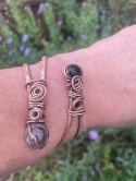 Autorska bransoletka miedziana pełnia mocy prezentująca naturalny kryształ górski oraz czarny onyks w technice wire wrapping trzymana w dłoni kobiety na tle ogrodu.